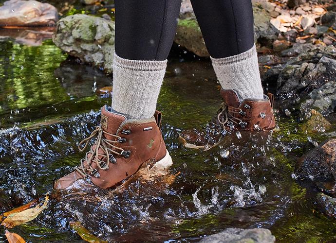 The image features a person walking in water wearing the Thatcher Women's Waterproof Sneakerboots in Toffee.