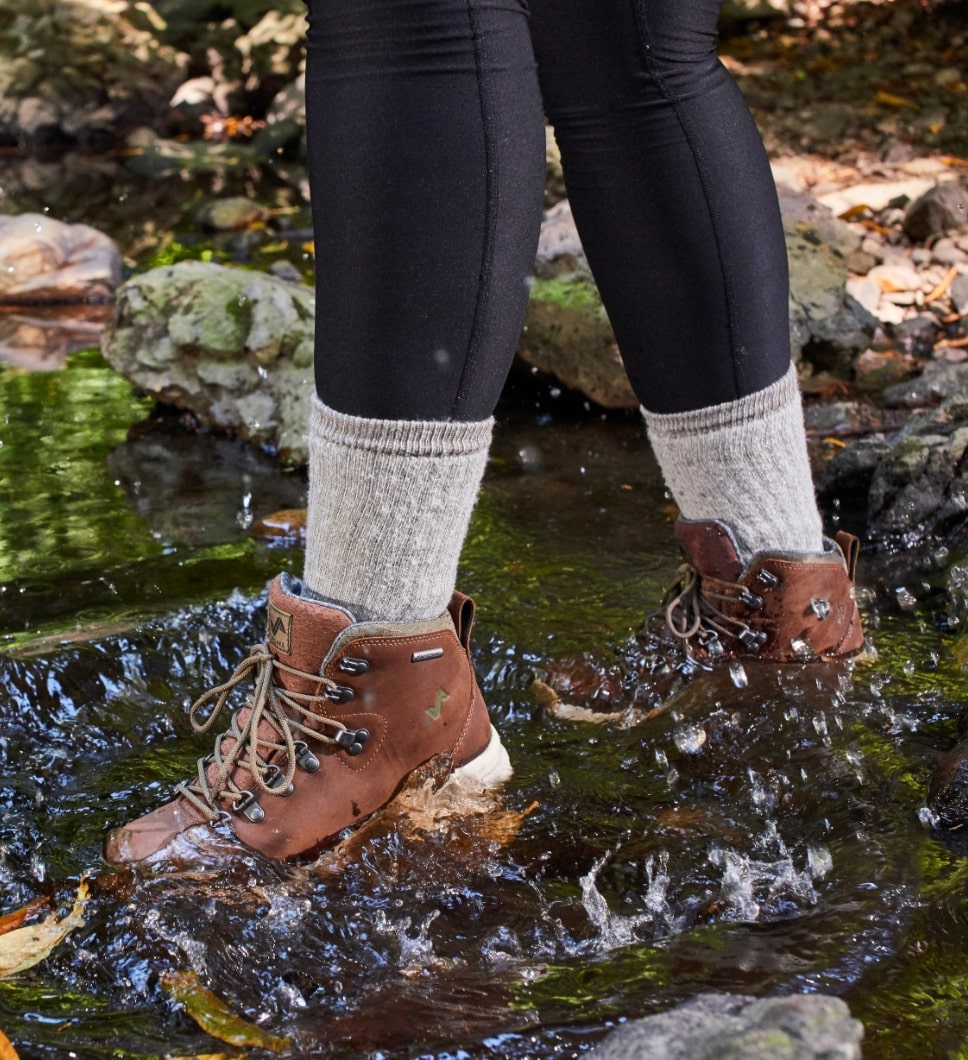 The image features a person walking in water wearing the Thatcher Women's Waterproof Sneakerboots in Toffee.